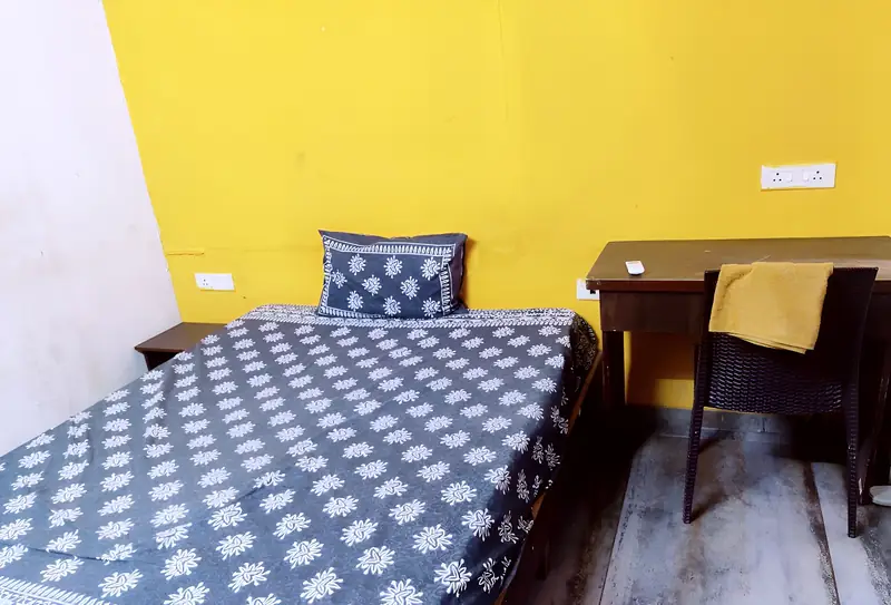 Single bed with printed bedspread, desk and chair against a yellow wall