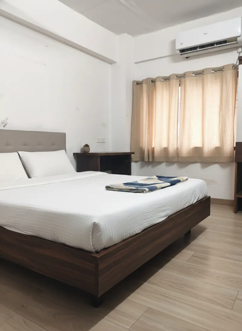 Room with one large bed in white linen with a padded headboard, a bedside table, a wall-mounted AC and beige curtains at the window